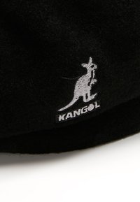 Sombrero de fieltro negro con un logotipo de canguro bordado en blanco y una etiqueta negra "KANGOL". Textura suave, forma redondeada, ala plana.
