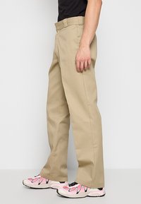 Beige kalhoty s rovnými nohavicemi a texturovaným povrchem, s plochým předním dílem a poutky na pásek, v kombinaci s růžovými a černými teniskami.