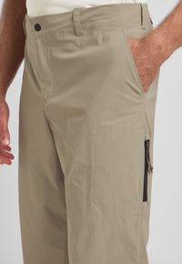 Pantaloni cargo beige in tessuto leggero con finitura testurizzata. Presentano tasca laterale con zip e tasche frontali standard con passanti per cintura.