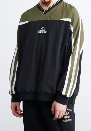 Mężczyzna noszący czarną i oliwkową bluzę Adidas z białymi paskami na rękawach oraz pasujące czarne spodnie Adidas z logo i napisem na udzie.