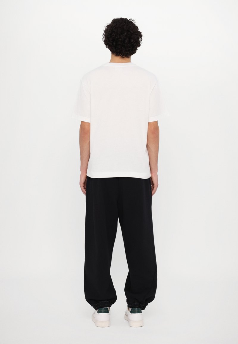 Personne aux cheveux bouclés, vue de dos, portant un t-shirt blanc uni, un pantalon noir ample et des baskets blanches avec des détails vert foncé.
