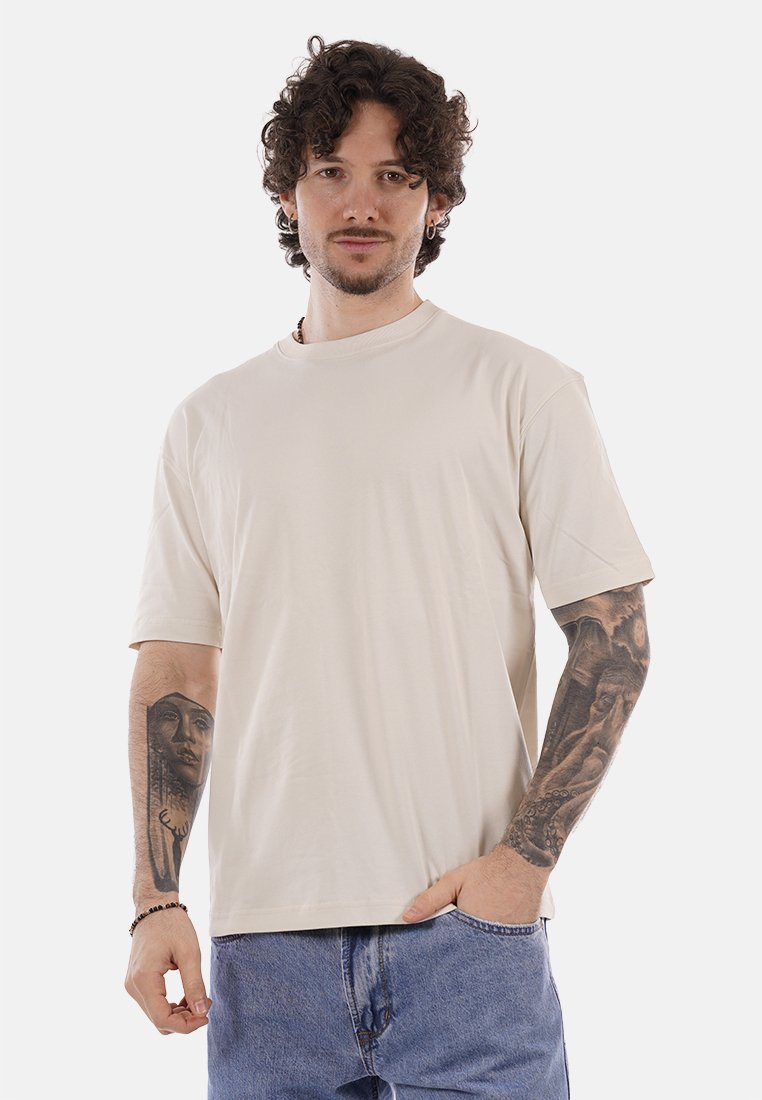 Homme aux cheveux bouclés et foncés portant un t-shirt beige et un jean bleu, montrant des tatouages sur les deux bras, debout devant un fond blanc uni.