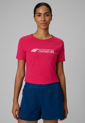 Kobieta o ciemnych włosach w jaskrawo różowym T-shircie z napisem „4F Outdoors” i granatowych szortach, stojąca na jednolitym szarym tle.