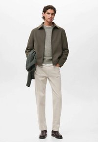 Giacca zip-in verde oliva sopra un maglione grigio chiaro, abbinata a pantaloni crema e stivali marroni scuri. Il modello tiene una sciarpa in maglia grigia.