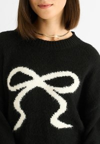 Maglione lavorato a maglia nero con un grande design di fiocco bianco sul davanti. La texture è morbida e soffice, con un classico collo rotondo.