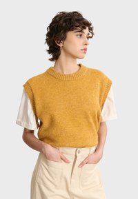 Senfgelbes ärmelloses Strickoberteil mit geripptem Halsausschnitt, über einem weißen kurzärmligen Hemd getragen; weiche Textur, lässige Passform.