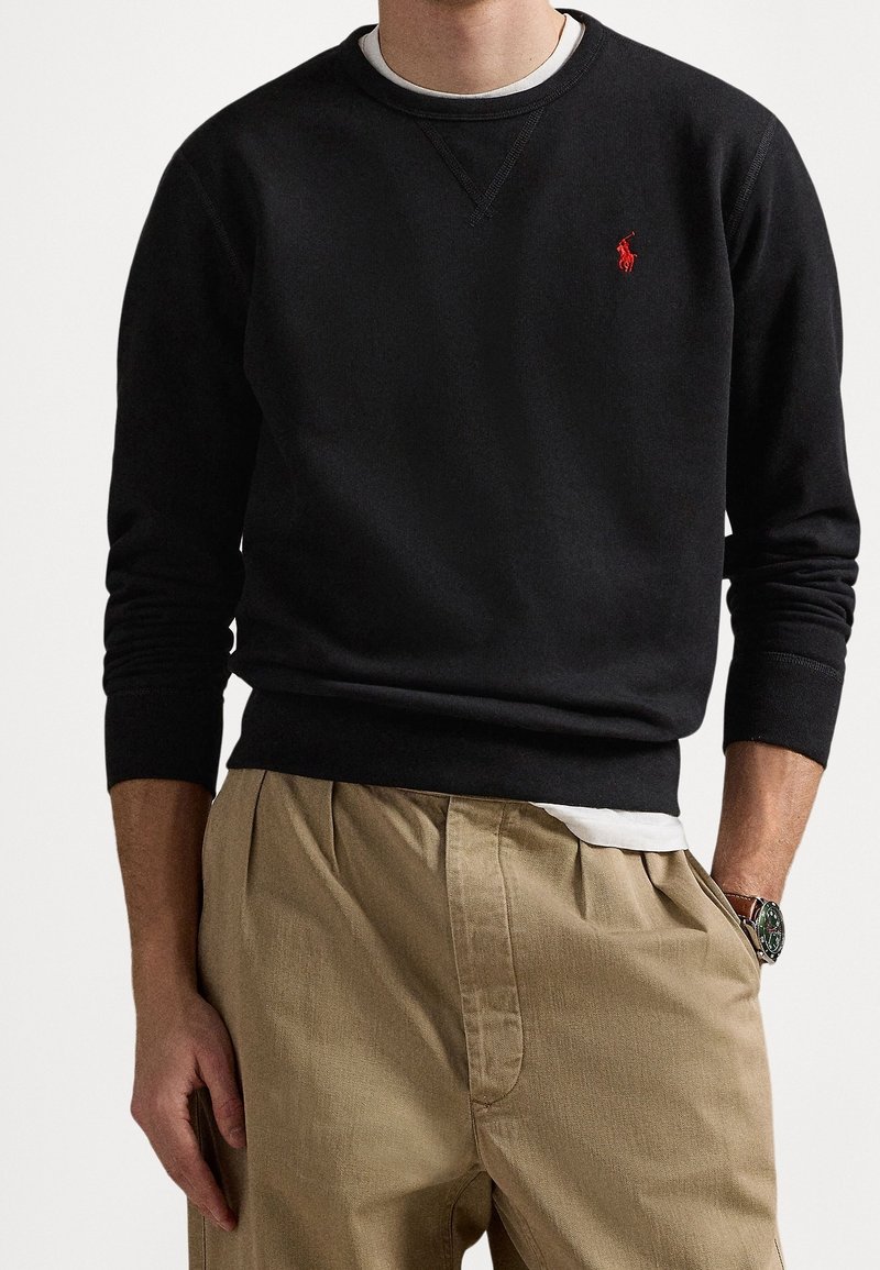 Homme portant un sweat-shirt noir à col ras du cou avec un logo rouge, un sous-pull blanc, un pantalon plissé beige et une montre-bracelet avec un bracelet sombre.