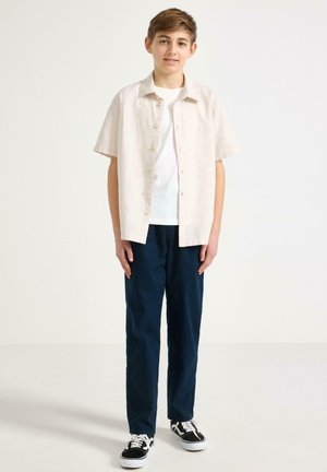 Teini-ikäinen poika seisoo, päällään beige lyhythihainen napillinen paita valkoisen t-paidan päällä, tummansiniset housut ja mustavalkoiset tennarit, neutraali tausta.