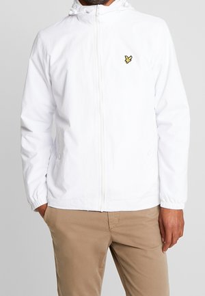 Mand iført en hvid lynlåstrøje med hætte og gul fugle-logo på brystet, kombineret med beige bukser, stående med hænderne i lommerne.