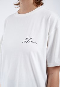 T-shirt blanc à manches courtes en coton doux, avec un logo noir brodé sur la poitrine gauche.