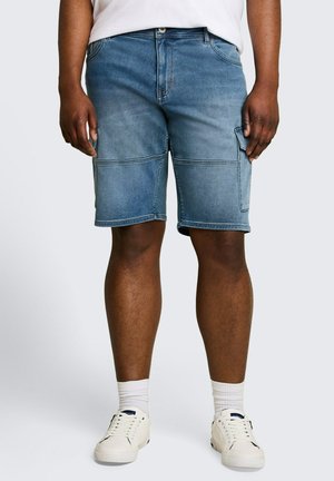 Mann trägt blaue Denim-Cargoshorts, weiße Socken und weiße Sneakers, steht vor einem schlichten hellen Hintergrund.