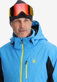 Giacca da sci blu con accenti neri, dotata di una cerniera gialla e un logo di ragno. Indossata con un berretto nero e occhiali protettivi riflettenti.