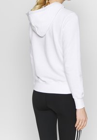 Felpa bianca con design aderente, tessuto testurizzato e cappuccio con coulisse, abbinata a leggings neri con tre strisce bianche.