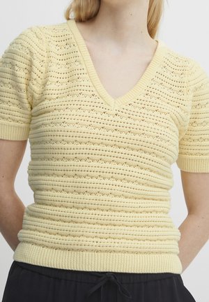 Vrouw draagt een lichtgeel kort mouw gebreid top met een V-hals en een getextureerd horizontaal patroon, gecombineerd met zwarte broek met trekkoord.