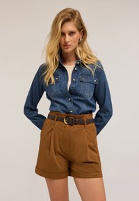 Camicia in denim con bottoni e due taschini sul petto, abbinata a pantaloni corti marroni a vita alta con cintura e design plissettato.