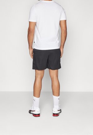 Maglietta bianca a maniche corte, pantaloncini sportivi neri, calzini bianchi con loghi e scarpe sportive con dettagli rossi. Outfit sportivo casual.