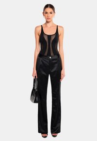 Svart mesh bodysuit med vertikala paneler, kombinerad med högmidjade svarta satinbyxor. Modellen håller en liten svart handväska.