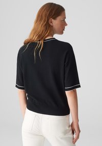 Schwarzer gestrickter Pullover mit kurzen Ärmeln, verziert mit weißen Abschlüssen an den Bündchen und am Ausschnitt, getragen mit weißen Hosen. Die Textur wirkt glatt.