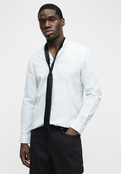 Νεαρός άνδρας που φοράει λευκό πουκάμισο με κουμπιά, με μια χαλαρά δεμένη μαύρη γραβάτα και μαύρο παντελόνι, στέκεται με το ένα χέρι στην τσέπη του.