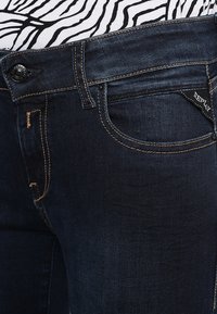 Jeans in denim blu scuro con un classico design a cinque tasche, dettagli di cucitura visibili e una piccola etichetta del marchio sulla tasca posteriore.