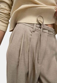 Beige Stoffhose mit Falten, verstellbarem Taillenzug und weichem Material, kombiniert mit einem hellbeigen gerippten Pullover.