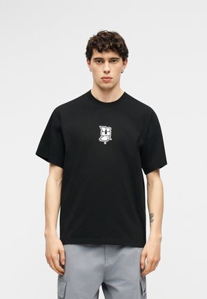 Νεαρός άνδρας μεσίου μήκους σγουρά μαλλιά, φορώντας μαύρο γραφικό μπλουζάκι και ανοιχτό γκρι παντελόνι τύπου cargo, στέκεται μπροστά σε ανοιχτό γκρι φόντο.