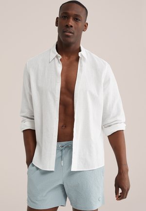 Mees, kes kannab valget nööpimata pika varrukaga särki, mille varrukad on üles keeratud, ja helesiniseid tekstuuriga seotava vööga lühikesi pükse.