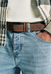 Ceinture marron en similicuir avec une boucle en métal noir, portée sur un jean en denim bleu clair, associée à une chemise beige et une veste chemise à carreaux.