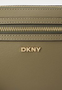 Olivenfarget DKNY-lommebok med gullglidelås og logo. Overflaten er teksturert med sømdetaljer, noe som gir et strukturert design.