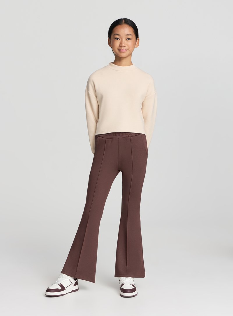 Felpa cropped beige a maniche lunghe abbinata a pantaloni marroni svasati. Completa l'outfit un paio di sneakers bianche con dettagli color granata.