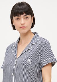 Navy och vit randig kortärmad pyjamaströja med en ficka som har en logotyp. Tyget verkar vara slätt med en klassisk kragdesign.