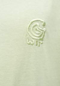 Camiseta de algodón verde claro con logo bordado en un diseño circular y letras texturizadas. Tejido suave con estética minimalista.