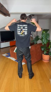 Homme portant un pantalon en cuir noir et un t-shirt noir avec des textes et des graphiques colorés, debout dans un salon avec des plantes et du mobilier.