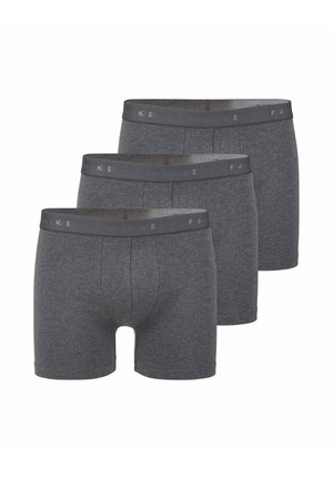 Uomo in piedi a piedi nudi con boxer grigio scuro su uno sfondo bianco semplice, rivolto in avanti con le braccia rilassate.