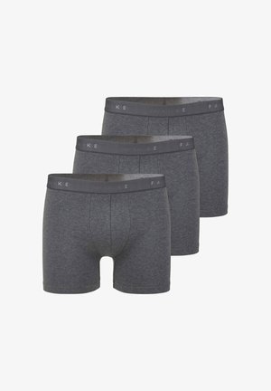 Mann steht barfuß in dunkelgrauen Boxershorts vor schlicht weißem Hintergrund, mit entspannten Armen nach vorne blickend.