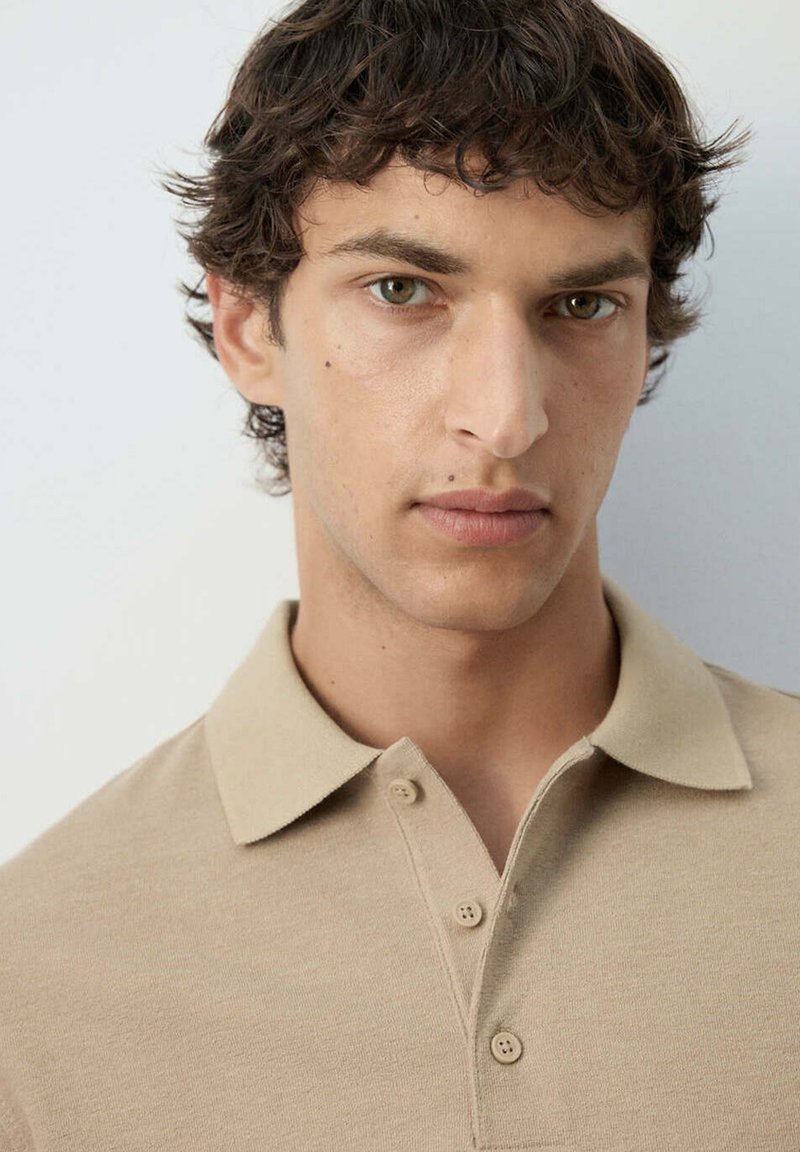 Joven con cabello corto, rizado y oscuro, que lleva una camisa beige con cuello y botones sobre un fondo liso y claro.