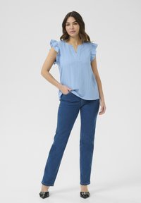 Top azzurro chiaro a maniche corte con dettagli arricciati e scollatura a V, abbinato a jeans dritti blu scuro e scarpe nere a punta.
