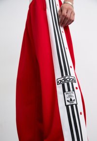 Sarkans sporta bikses ar baltu sānu paneļa, kurā ir trīs melnas svītras un Adidas logo uzšuvums, ar pogu aizdari gar sānu.