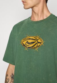 Grøn bomulds t-shirt med en slidteknik, der har et stort gult og sort Nike-logo omgivet af et tornlignende design på forsiden.
