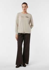 Beżowy sweter z metalicznym logo "LESS IS MORE", w połączeniu z ciemnobrązowymi spodniami typu dzwony. Sweter ma ściągacz u dołu i długie rękawy.