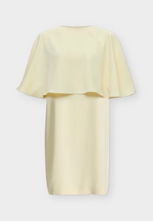 Donna bionda che indossa un vestito corto crema con maniche ampie, tiene in mano una grande pochette nera con frange e sandali neri con tacco.