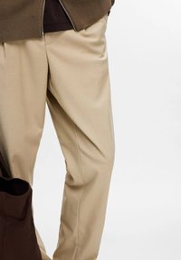 Pantalones beige ajustados con textura suave y diseño con pliegues, que cuentan con bolsillos laterales y un ligero brillo, combinados con un bolso de hombro oscuro.