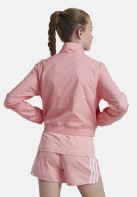Chaqueta cortavientos rosa con un cuello alto, puños elásticos y cinturilla. Presenta franjas deportivas blancas en las mangas, combinadas con pantalones cortos a juego.