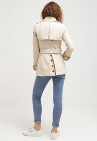 Trench-coat beige avec une taille ceinturée, des détails de boutons au dos et un col replié, associé à un jean bleu clair et des baskets blanches.