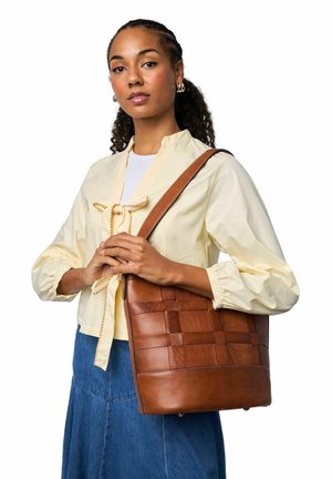 Braune geflochtene Ledertasche mit glatter Textur, flacher Form und langem Schulterriemen, gehalten von einer Person in einer hellgelben Bluse.