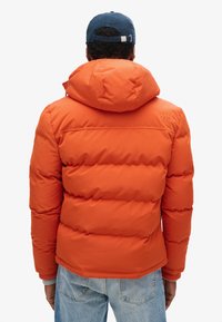 Chaqueta de plumas de color naranja brillante con capucha, que presenta secciones acolchadas horizontales y detalle del logotipo en el hombro. Combinada con jeans azules.