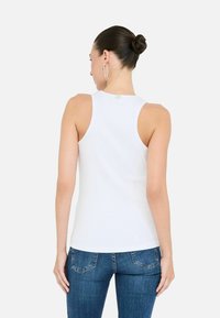 Model met de rug naar de camera, in een fitted wit tanktop en blauwe jeans, met een strakke bun en ronde oorbellen. Witte achtergrond.