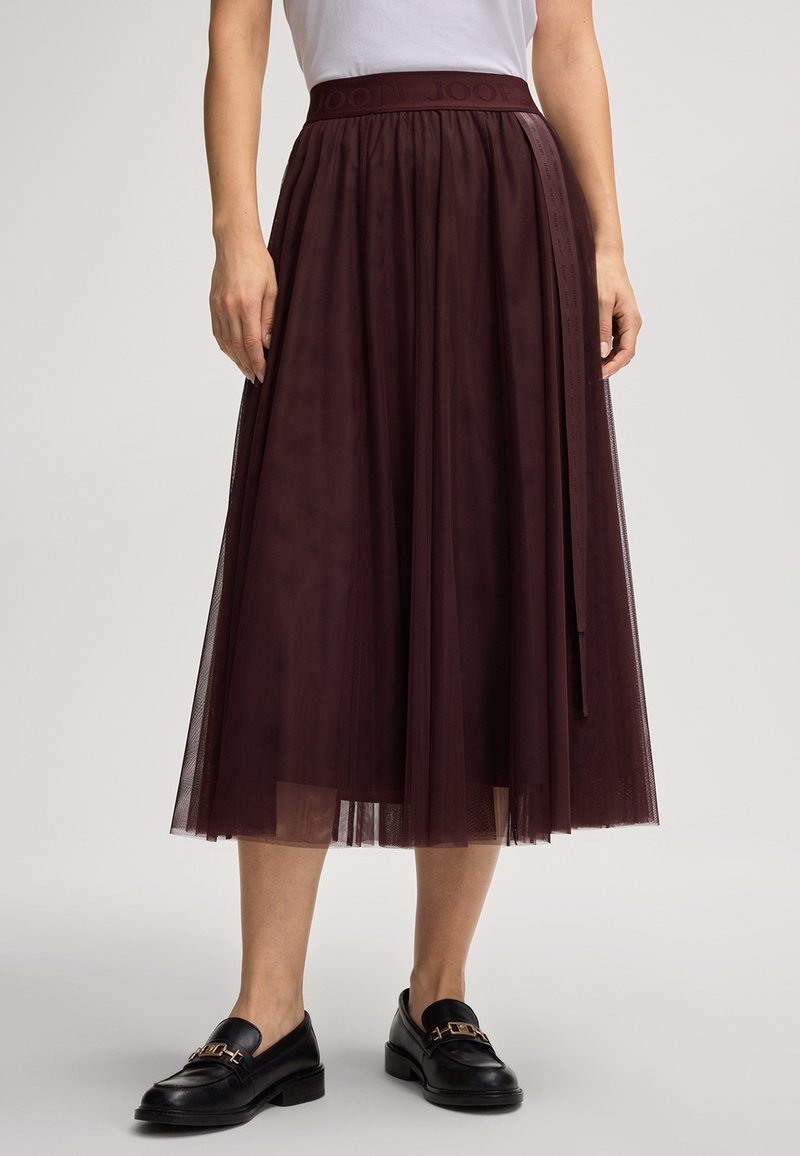 Woman wearing a burgundy tulle midi skirt with a branded waistband and black leather loafers with gold buckles.