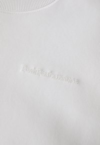 Felpa bianca in tessuto texturizzato con colletto a coste e logo "PeakPerformance" in rilievo sulla parte frontale. Finitura liscia.