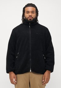Mand med dreadlocks, iført sort fleece-jakke med lynlås og beige bukser, står foran en ensfarvet hvid baggrund.